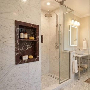 May include: A luxurious bathroom with marble walls and a glass shower enclosure. A recessed shelf with toiletries is visible. The shower features a rain shower head and a chrome faucet. The floor is tiled with white hexagonal tiles.