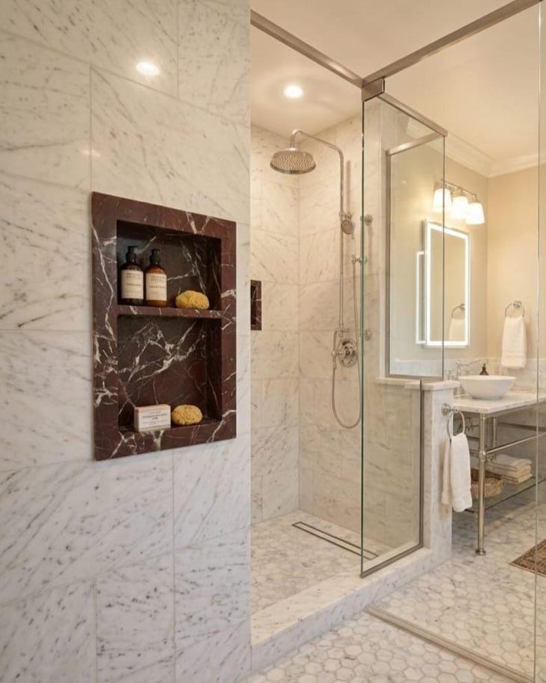 May include: A luxurious bathroom with marble walls and a glass shower enclosure. A recessed shelf with toiletries is visible. The shower features a rain shower head and a chrome faucet. The floor is tiled with white hexagonal tiles.