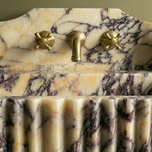 Pu&ograve; includere: Un lavabo in marmo con un motivo venato color crema e viola. Il lavabo ha un fronte scanalato e un alzatina decorativa. Sono visibili rubinetti color oro e un dosatore di sapone nero.