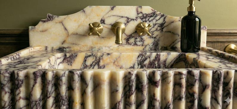 Pu&ograve; includere: Un lavabo in marmo con un motivo venato color crema e viola. Il lavabo ha un fronte scanalato e un alzatina decorativa. Sono visibili rubinetti color oro e un dosatore di sapone nero.