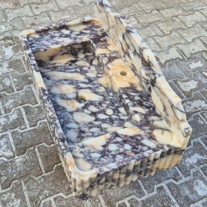 Pu&ograve; includere: Un lavandino rettangolare in materiale color crema con venature viola scuro e grigie. Il lavandino ha un ripiano integrato e un foro di scarico. L'esterno presenta un design a coste. Il lavandino &egrave; posto su una superficie di mattoni grigi.