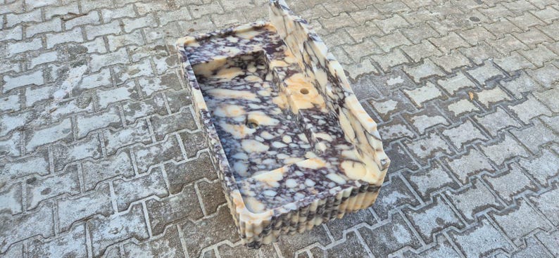 Pu&ograve; includere: Un lavandino rettangolare in materiale color crema con venature viola scuro e grigie. Il lavandino ha un ripiano integrato e un foro di scarico. L'esterno presenta un design a coste. Il lavandino &egrave; posto su una superficie di mattoni grigi.