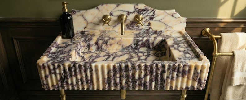 Pu&ograve; includere: Un lavabo da bagno in marmo con un motivo venato color crema e viola. Il lavabo ha un fronte scanalato e finiture dorate, tra cui un rubinetto e due maniglie. Un dispenser di sapone nero &egrave; appoggiato sul bordo del lavabo. Un portasciugamani dorato &egrave; visibile a destra.