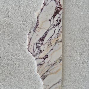 Puede incluir: Una losa rectangular de m&aacute;rmol blanco con vetas irregulares y prominentes en color p&uacute;rpura oscuro y dorado. El lado izquierdo de la losa de piedra tiene una forma recortada org&aacute;nica y dentada, mientras que el lado derecho es recto y vertical.