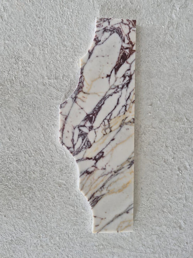 Puede incluir: Una losa rectangular de m&aacute;rmol blanco con vetas irregulares y prominentes en color p&uacute;rpura oscuro y dorado. El lado izquierdo de la losa de piedra tiene una forma recortada org&aacute;nica y dentada, mientras que el lado derecho es recto y vertical.