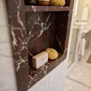 May include: A built-in marble shelf in a bathroom, featuring a deep red and white veined pattern. The shelf holds two bottles of shampoo, a natural sponge, and a bar of soap labeled "ANYA ROSE GERANIUM".
