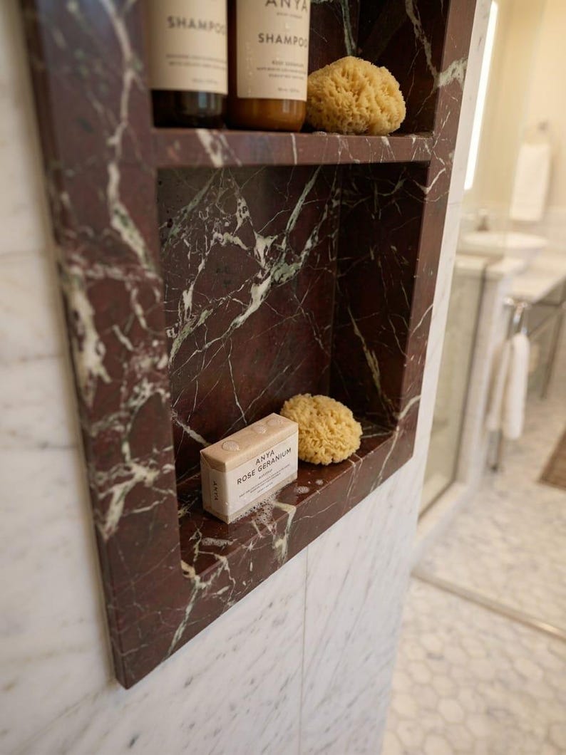 May include: A built-in marble shelf in a bathroom, featuring a deep red and white veined pattern. The shelf holds two bottles of shampoo, a natural sponge, and a bar of soap labeled "ANYA ROSE GERANIUM".