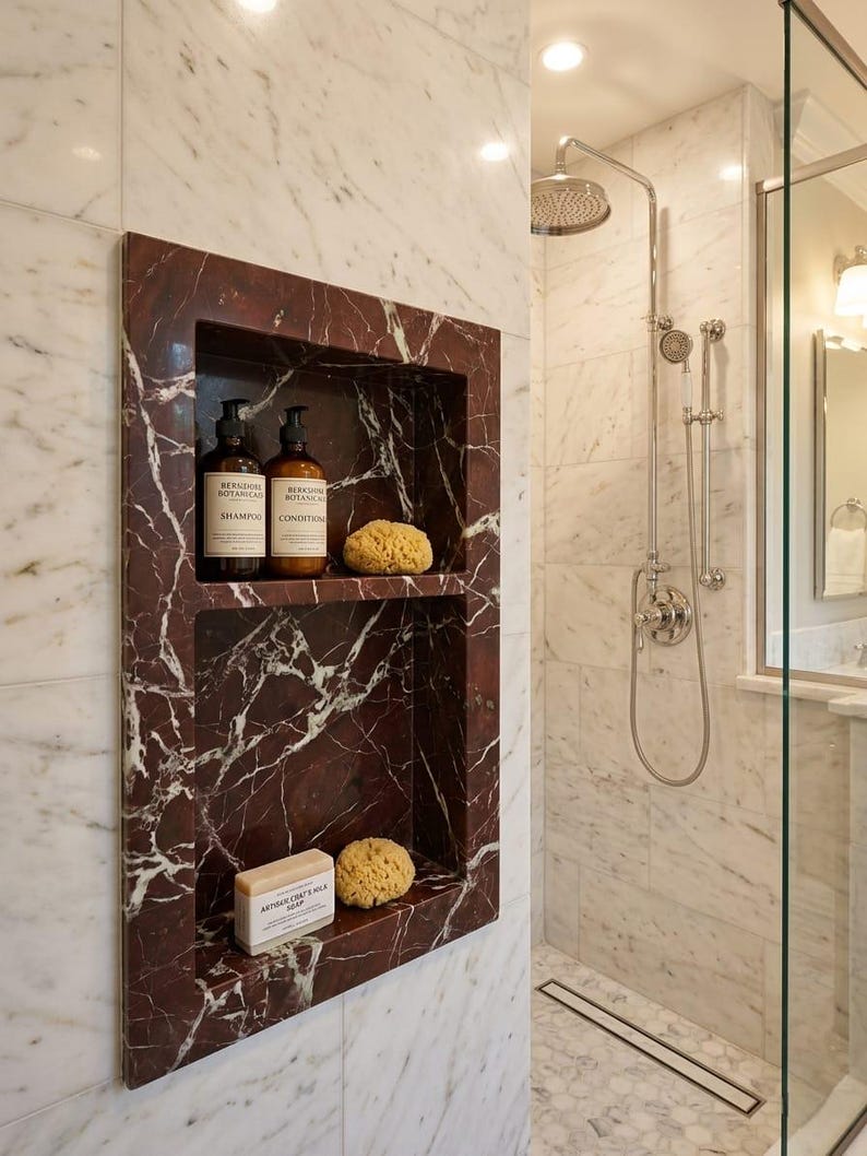 May include: A built-in shower niche with a dark red marble finish, holding shampoo, conditioner, a bar of soap, and a natural sponge. The shower features a rainfall showerhead and a glass enclosure, set against a backdrop of white marble tiles.