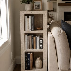 Pu&ograve; includere: Una libreria rettangolare in pietra beige porosa con tre ripiani aperti. I ripiani contengono vari libri con titoli tra cui Century, Vanity Fair 100 Years e Picaso, insieme a una piccola ciotola di legno e un vaso in ceramica beige.