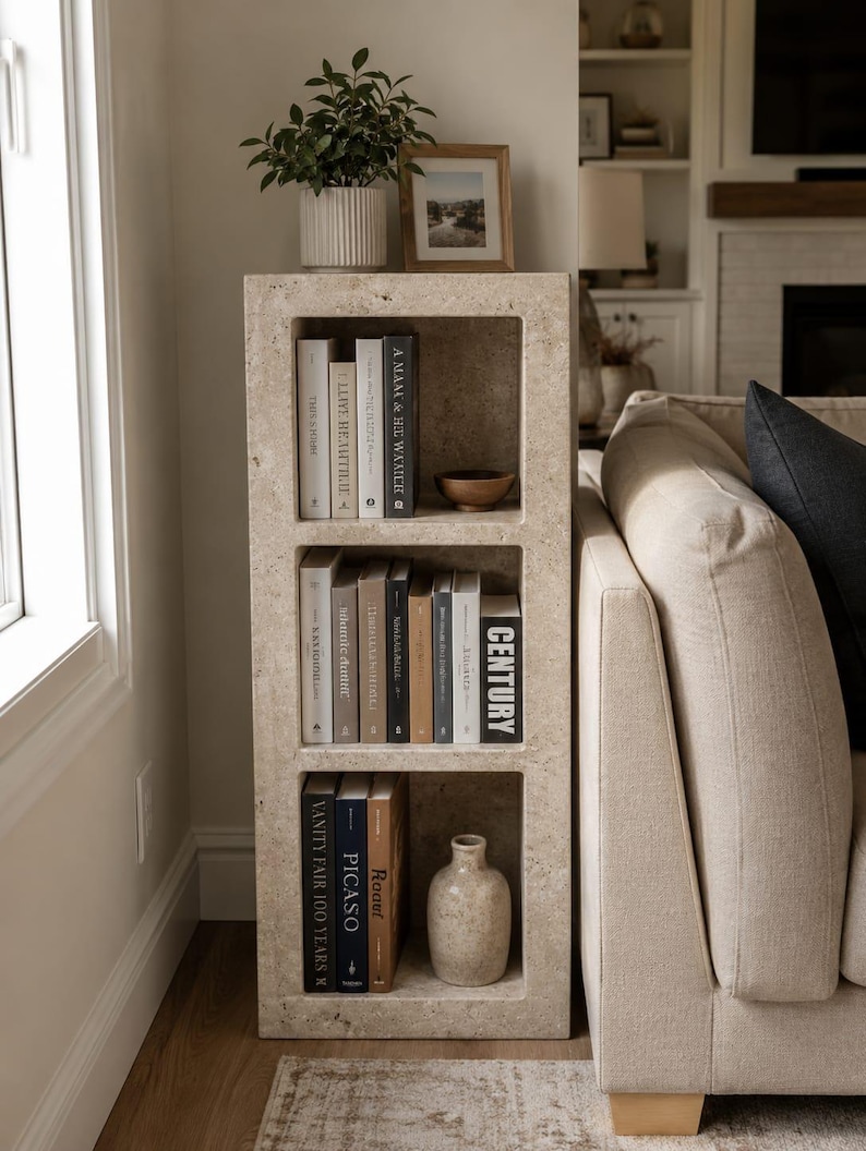 Pu&ograve; includere: Una libreria rettangolare in pietra beige porosa con tre ripiani aperti. I ripiani contengono vari libri con titoli tra cui Century, Vanity Fair 100 Years e Picaso, insieme a una piccola ciotola di legno e un vaso in ceramica beige.