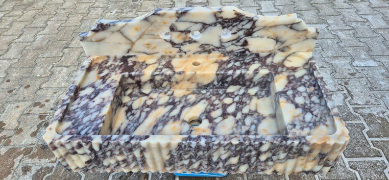 Pu&ograve; includere: Un lavabo rettangolare in marmo con alzatina. Il lavabo presenta un motivo a crema, viola e marrone. Il lavabo ha uno scarico centrale e due fori per il rubinetto. I bordi del lavabo sono smerlati.