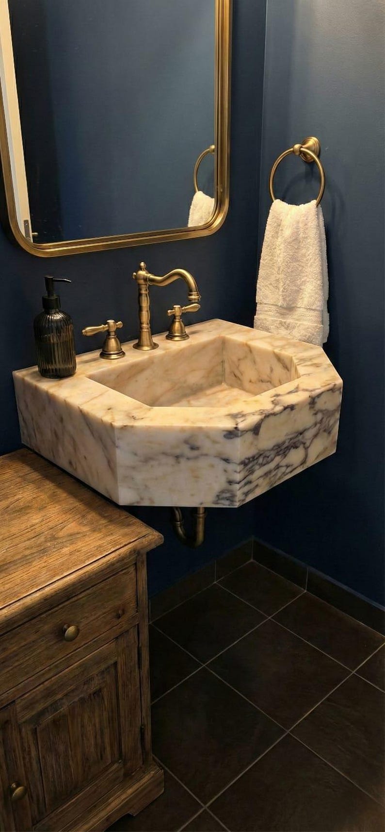 May include: An octagonal marble sink with brass taps and a matching mirror. A wooden cabinet sits below the sink. A white towel hangs on a brass ring. The sink has a cream and grey pattern.