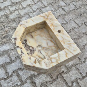 May include: A marble sink with a unique geometric design. The sink features a rectangular basin with a rounded drain and a faucet hole. The marble has a cream and brown veining pattern. The sink is set on a grey brick surface.