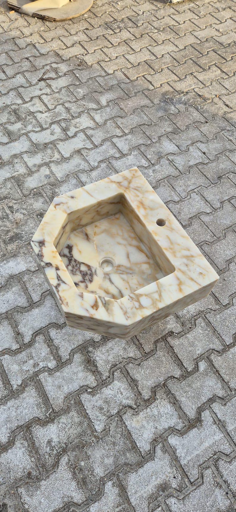 May include: A marble sink with a unique geometric design. The sink features a rectangular basin with a rounded drain and a faucet hole. The marble has a cream and brown veining pattern. The sink is set on a grey brick surface.