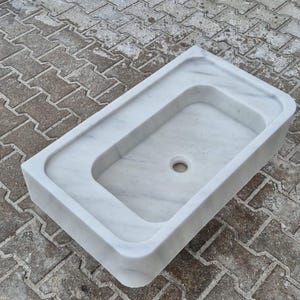 May include: A rectangular, white marble sink with a rounded interior basin and a central drain hole. The sink has a raised border and is set against a textured, gray surface.