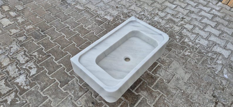 May include: A rectangular, white marble sink with a rounded interior basin and a central drain hole. The sink has a raised border and is set against a textured, gray surface.