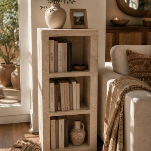 Pu&ograve; includere: Una libreria rettangolare in pietra beige chiaro con tre scomparti aperti. I ripiani contengono vari libri con dorsi di colori neutri, una piccola ciotola scura e un vaso in ceramica testurizzato. La struttura presenta una finitura in pietra naturale porosa.