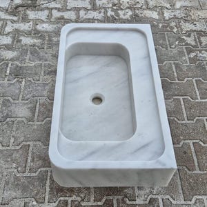 May include: A white marble sink with a rectangular basin and a rounded edge. The sink has a central drain hole and is set against a textured, gray brick surface. The sink's design is minimalist and modern.