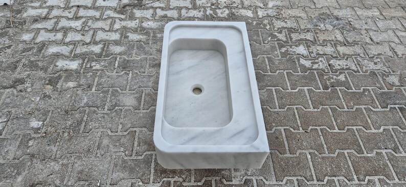 May include: A white marble sink with a rectangular basin and a rounded edge. The sink has a central drain hole and is set against a textured, gray brick surface. The sink's design is minimalist and modern.