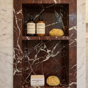 May include: A built-in shower niche with a deep red marble finish. The top shelf holds two brown bottles of shampoo and conditioner, and a natural sponge. The bottom shelf has a bar of soap and another sponge.