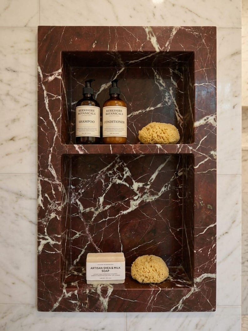 May include: A built-in shower niche with a deep red marble finish. The top shelf holds two brown bottles of shampoo and conditioner, and a natural sponge. The bottom shelf has a bar of soap and another sponge.