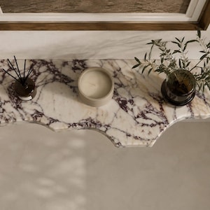 Puede incluir: Un estante flotante de pared hecho de m&aacute;rmol blanco con vetas p&uacute;rpuras prominentes y un borde org&aacute;nico irregular. El estante sostiene un peque&ntilde;o difusor de varillas de vidrio con palitos negros, una vela redonda beige y un florero de vidrio con follaje verde.