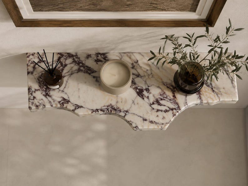 Puede incluir: Un estante flotante de pared hecho de m&aacute;rmol blanco con vetas p&uacute;rpuras prominentes y un borde org&aacute;nico irregular. El estante sostiene un peque&ntilde;o difusor de varillas de vidrio con palitos negros, una vela redonda beige y un florero de vidrio con follaje verde.