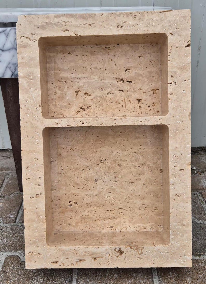 May include: A rectangular, beige stone tray with two recessed compartments. The stone has a porous texture and a natural, earthy appearance. The tray is likely for decorative or functional use, such as holding soap or small items.