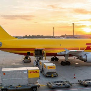 Puede incluir: Un avi&oacute;n de carga amarillo brillante con la marca DHL en rojo en el fuselaje y la cola, estacionado en la pista de un aeropuerto durante el atardecer. Varios contenedores de carga met&aacute;licos con logos DHL est&aacute;n cerca de la bodega abierta.