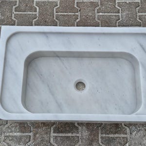 May include: A rectangular white marble sink with a rounded basin and a central drain hole. The sink has a smooth, polished surface with subtle gray veining, and a raised border.