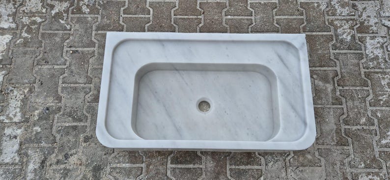 May include: A rectangular white marble sink with a rounded basin and a central drain hole. The sink has a smooth, polished surface with subtle gray veining, and a raised border.