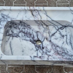May include: Rectangular marble sink with a white base and dark gray veining. The sink has a rounded interior basin and a small drain hole. The sink is made of stone and is suitable for a bathroom.