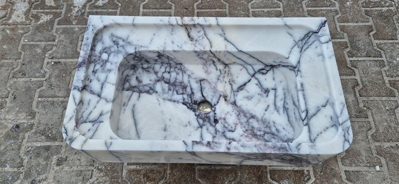 May include: Rectangular marble sink with a white base and dark gray veining. The sink has a rounded interior basin and a small drain hole. The sink is made of stone and is suitable for a bathroom.