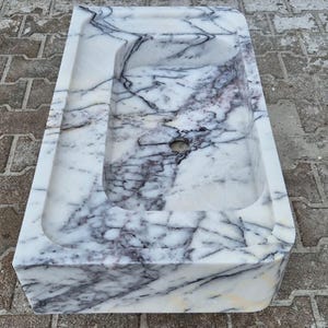 May include: A rectangular marble sink with a basin and a drain hole. The sink is white with dark gray veining throughout. The sink is sitting on a gray and brown brick surface.