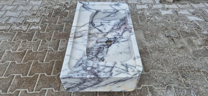 May include: A rectangular marble sink with a basin and a drain hole. The sink is white with dark gray veining throughout. The sink is sitting on a gray and brown brick surface.