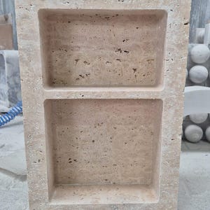 May include: A rectangular, beige stone shelf with two recessed compartments. The stone has a textured surface with small holes and variations in color. The shelf is likely for displaying items or storage.