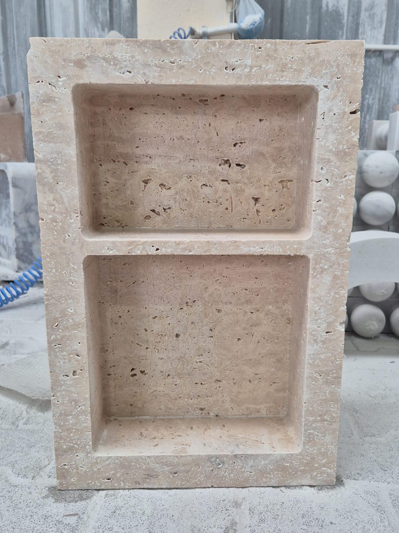 May include: A rectangular, beige stone shelf with two recessed compartments. The stone has a textured surface with small holes and variations in color. The shelf is likely for displaying items or storage.