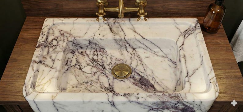 May include: A rectangular marble sink with a white and purple veined pattern. The sink has a brass drain cover and faucet. It is set on a wooden countertop with a brown glass bottle.