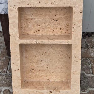 May include: A rectangular, beige stone shelf with two recessed compartments. The stone has a natural, textured appearance with small holes and variations in color. The shelf is designed for storage or display and is likely for a bathroom or kitchen.