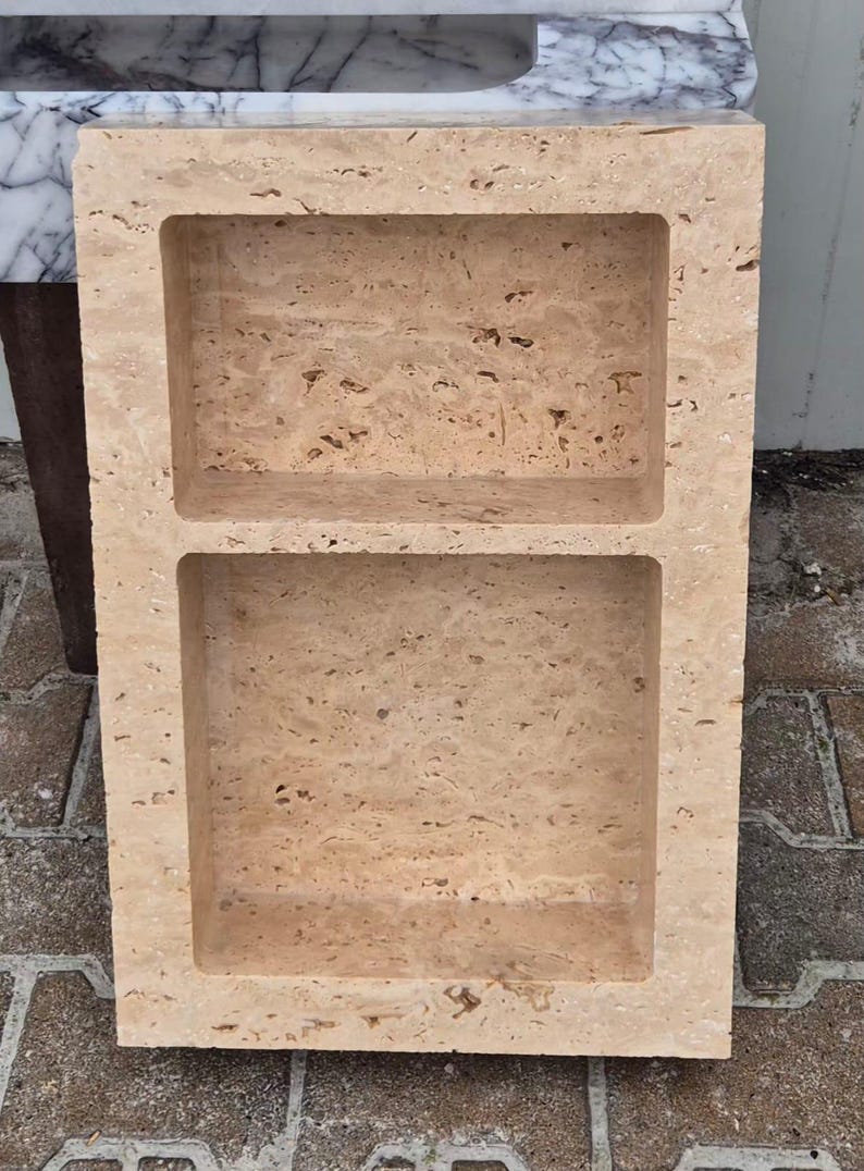 May include: A rectangular, beige stone shelf with two recessed compartments. The stone has a natural, textured appearance with small holes and variations in color. The shelf is designed for storage or display and is likely for a bathroom or kitchen.