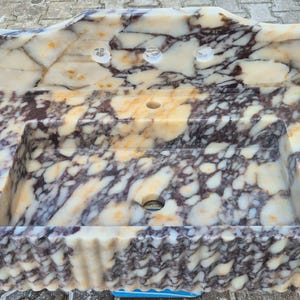 Pu&ograve; includere: Un lavabo rettangolare in marmo con un motivo a crema, marrone e viola. Il lavabo ha un alzatina con due fori per rubinetti e uno scarico centrale. Il bordo anteriore ha un design decorativo smerlato.