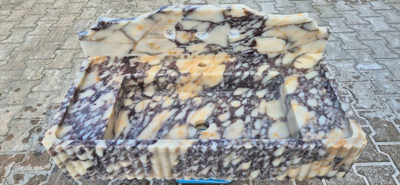 Pu&ograve; includere: Un lavabo rettangolare in marmo con un motivo a crema, marrone e viola. Il lavabo ha un alzatina con due fori per rubinetti e uno scarico centrale. Il bordo anteriore ha un design decorativo smerlato.