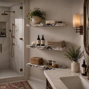 May include: Three floating shelves with a black and white marble pattern mounted on a wall. The shelves hold a potted plant, folded beige towels, two amber glass pump bottles, a candle, a woven basket, and small glass jars with metal lids.