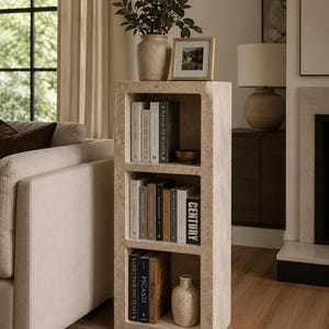 Pu&ograve; includere: Una libreria rettangolare in pietra travertino beige con tre ripiani aperti. Il ripiano superiore ospita una piccola pianta in un vaso, una cornice portafoto e dei libri. Il ripiano centrale contiene dei libri, quello inferiore libri e un vaso.