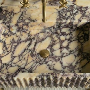 Pu&ograve; includere: Un lavabo rettangolare in marmo con un motivo venato color crema e viola. Il lavabo ha un rubinetto e uno scarico in ottone e un bordo smerlato decorativo. Una bottiglia scura poggia sul lato.