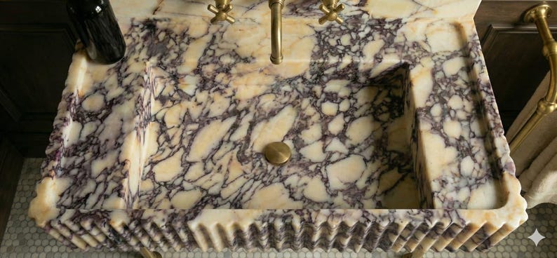 Pu&ograve; includere: Un lavabo rettangolare in marmo con un motivo venato color crema e viola. Il lavabo ha un rubinetto e uno scarico in ottone e un bordo smerlato decorativo. Una bottiglia scura poggia sul lato.