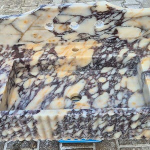 Pu&ograve; includere: Un lavandino rettangolare in marmo con un motivo a crema, marrone e viola. Il lavandino ha un alzatina integrata con tre fori per rubinetti e uno scarico centrale. I bordi hanno un design ondulato e strutturato.