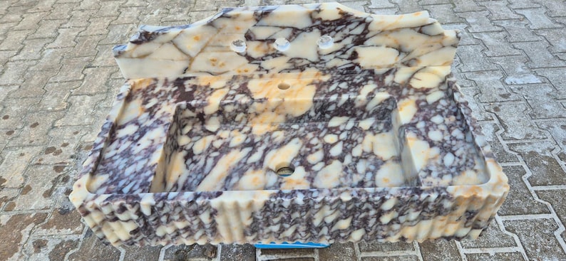 Pu&ograve; includere: Un lavandino rettangolare in marmo con un motivo a crema, marrone e viola. Il lavandino ha un alzatina integrata con tre fori per rubinetti e uno scarico centrale. I bordi hanno un design ondulato e strutturato.