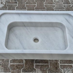May include: A rectangular white marble sink with a rounded interior basin and a central drain hole. The sink has a smooth, polished surface and is designed for bathroom use.