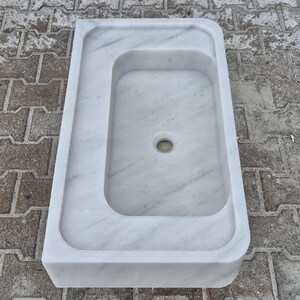 May include: A white marble sink with a rectangular basin and a smaller, offset rectangular area. The sink has a rounded edge and a drain hole in the center of the basin. The sink is made of white marble with gray veining.